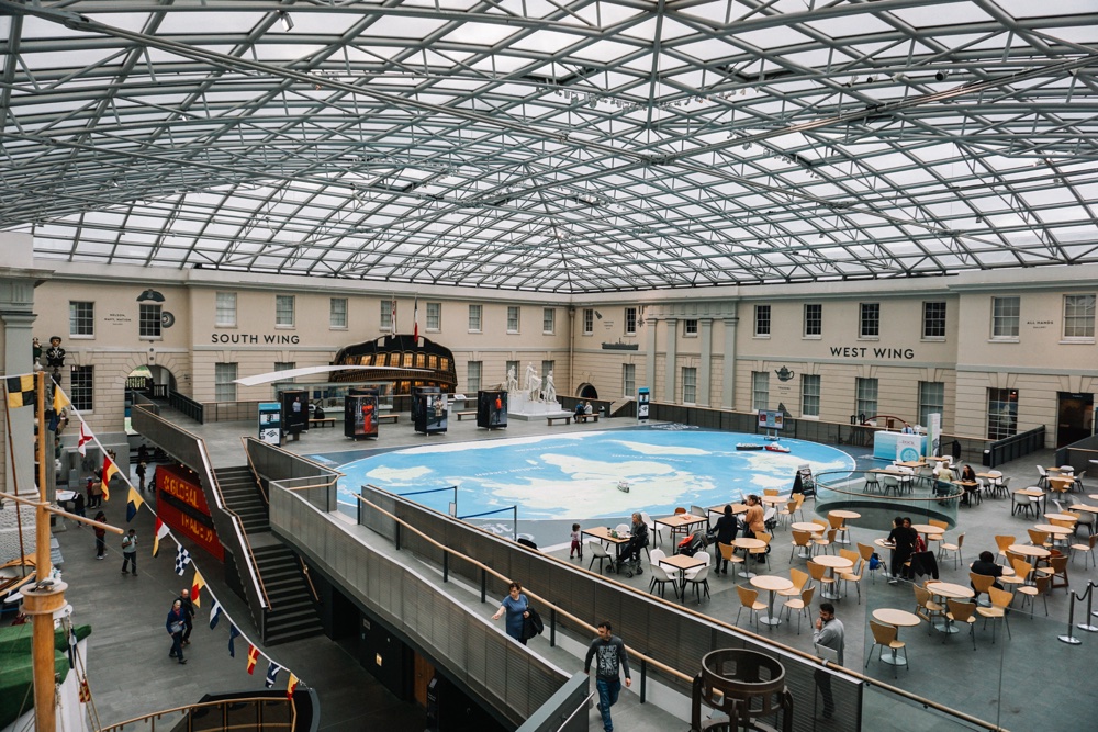 A spacious museum atrium featuring a large world map, dining tables, and visitors exploring exhibits under a glass roof.