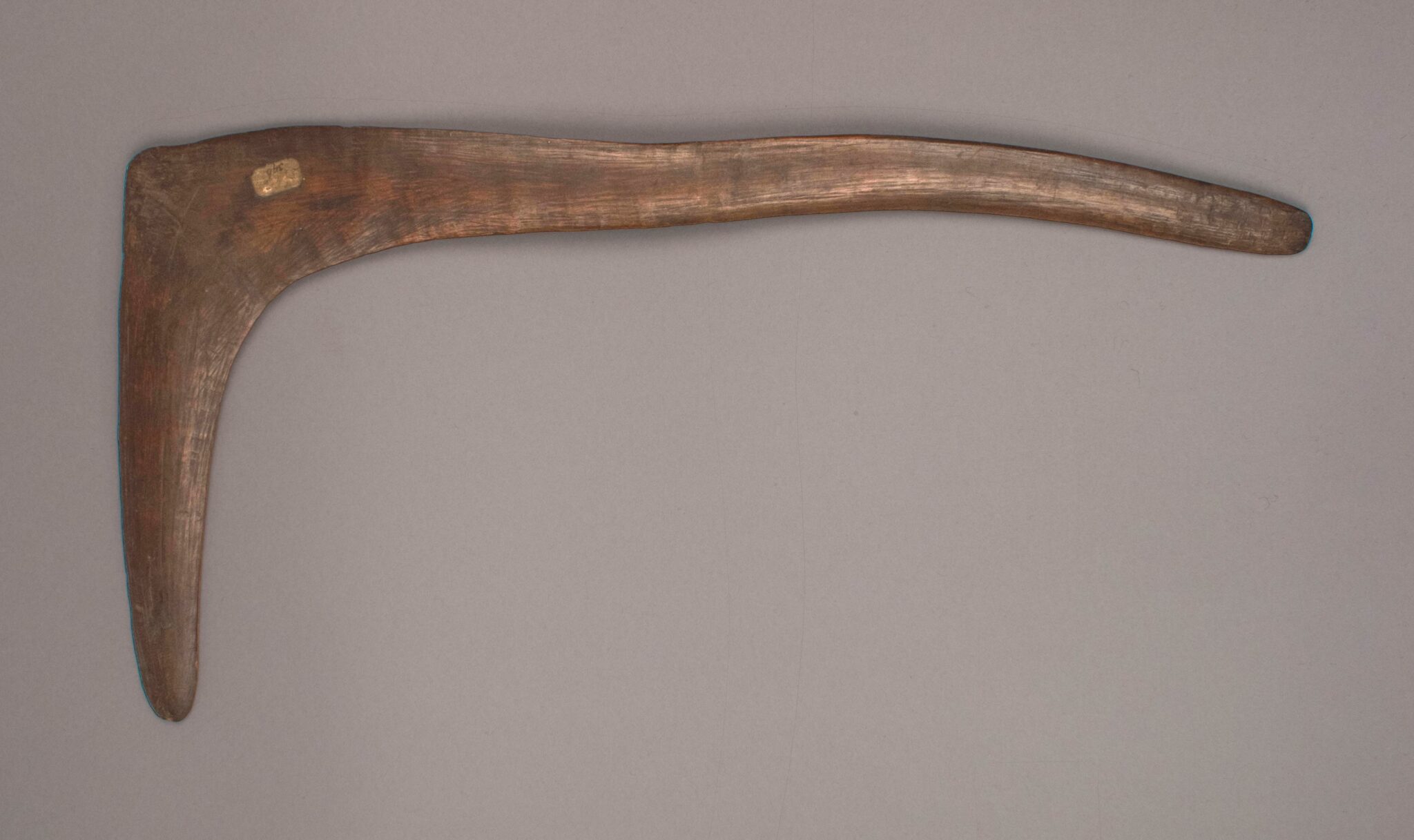 A worn wooden object in a L shape, showing distinct grains and an antique label, set against a neutral backdrop.