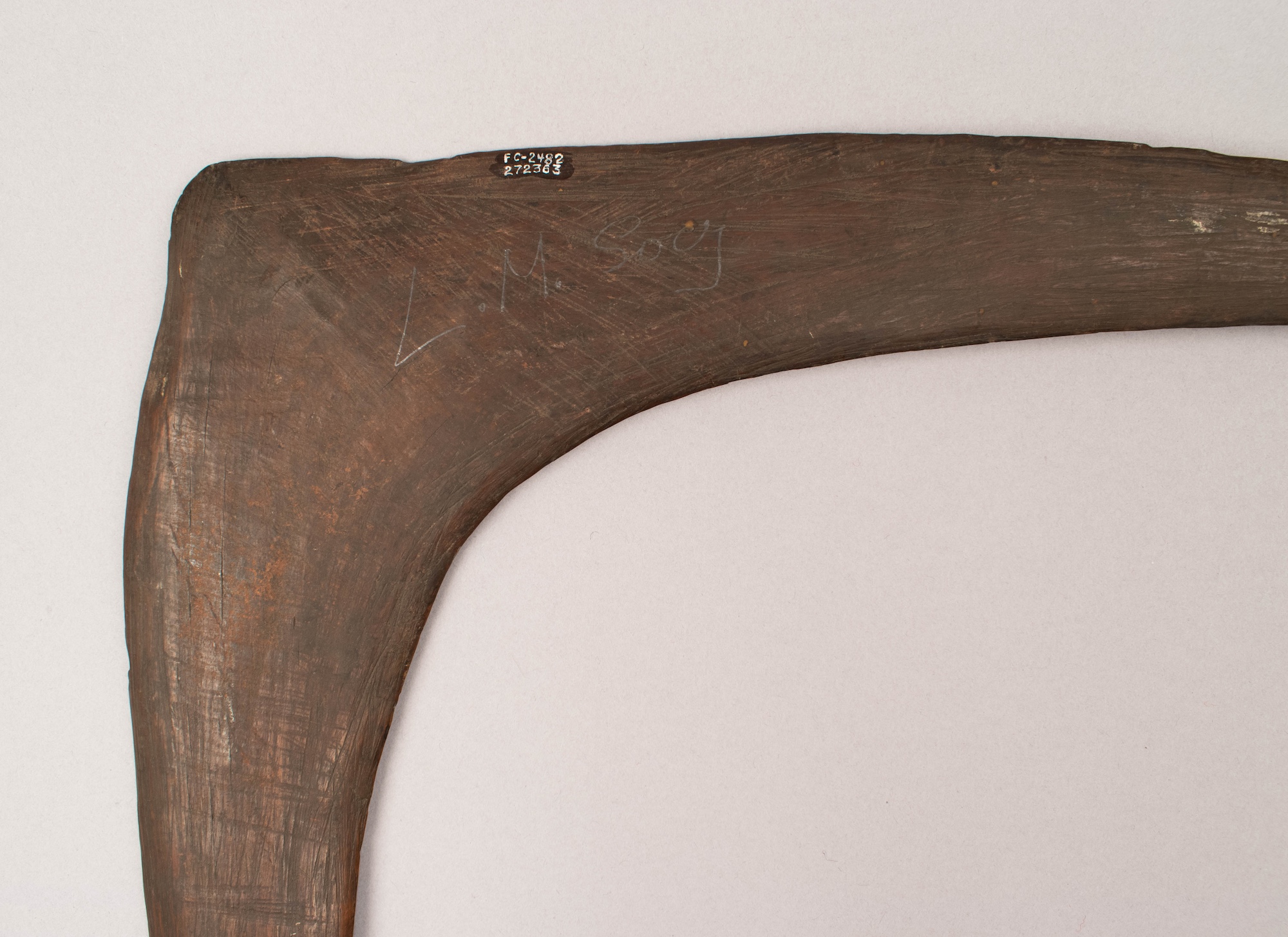 Close-up of a large, curved wooden artifact, featuring etched markings and a tag with numbers, displayed against a light background.