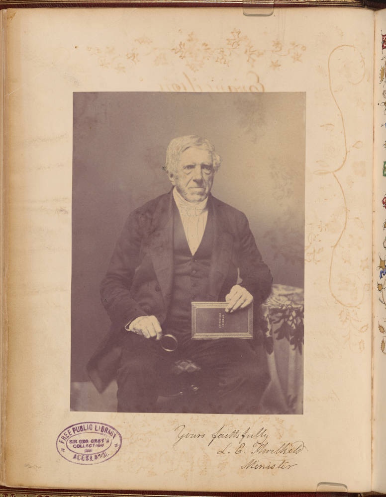 A seated man in 19th-century attire holds a book and a small object, surrounded by a floral-patterned tablecloth.