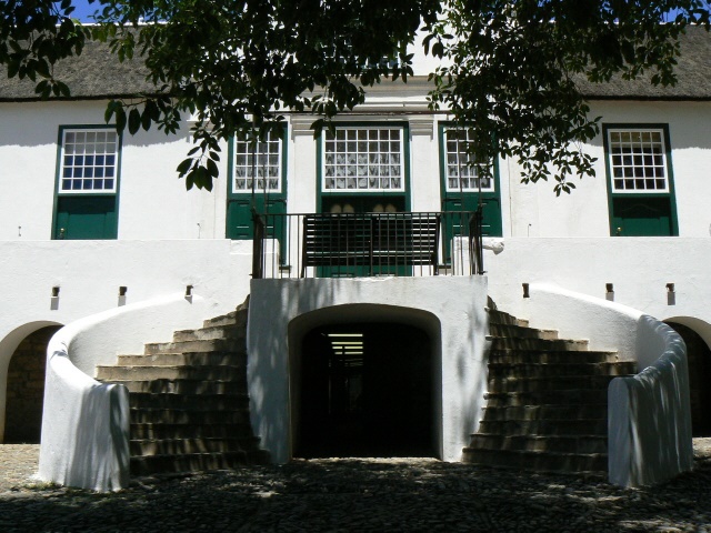 White colonial-style building with green doors, featuring a grand staircase and an arched entrance framed by trees.