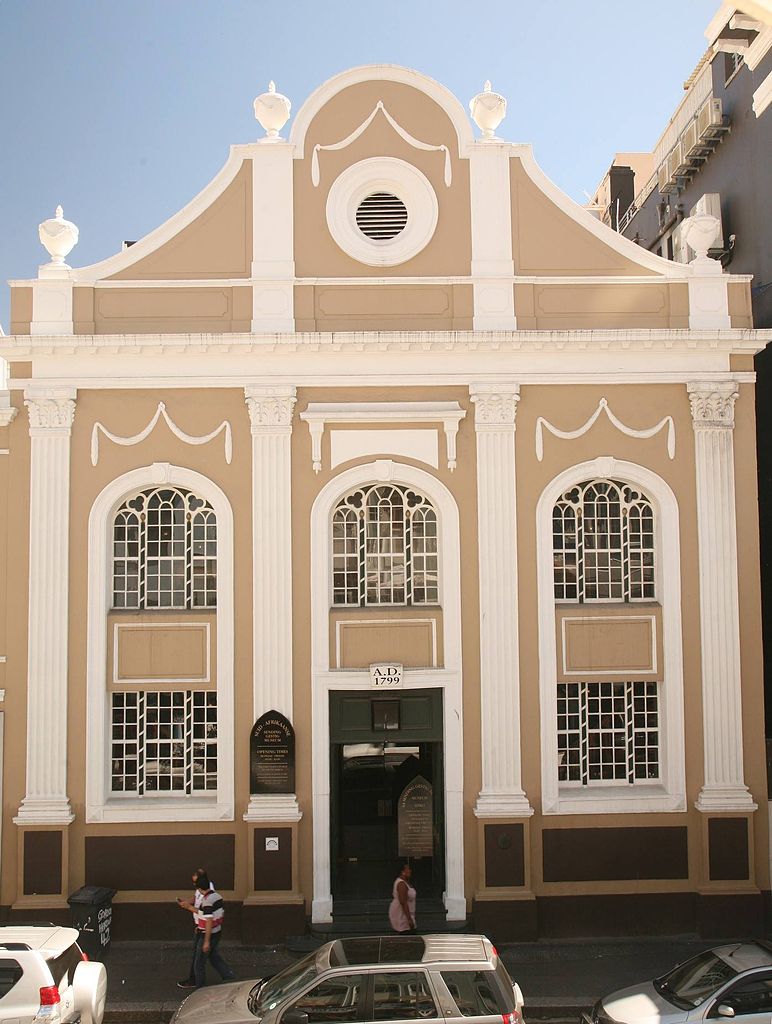 Historic building with a beige facade, prominent columns, arched windows, and a decorative gable, featuring people walking by.