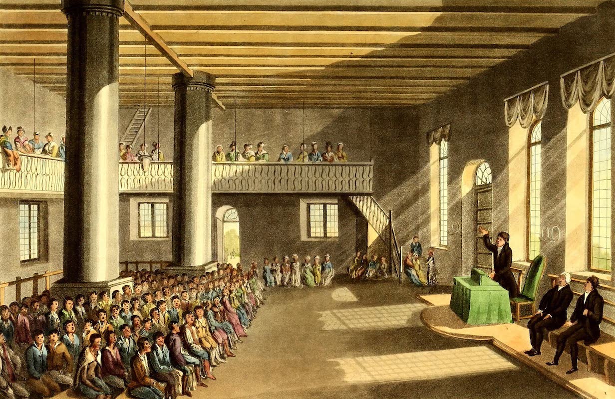 A large audience attentively listens to a speaker at a historic hall, with a balcony above and sunlight streaming through tall windows.