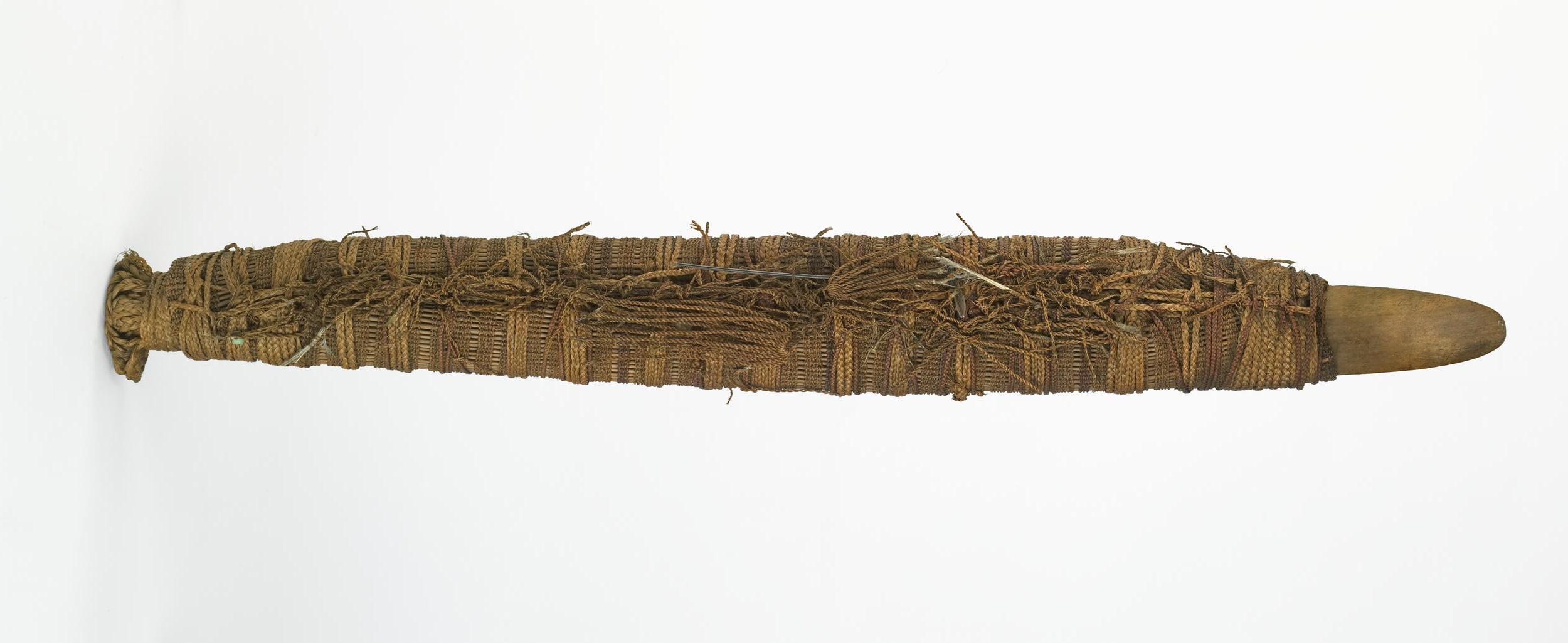 A long, woven ceremonial object with a pointed wooden end, intricately wrapped in natural fibers and plant materials.