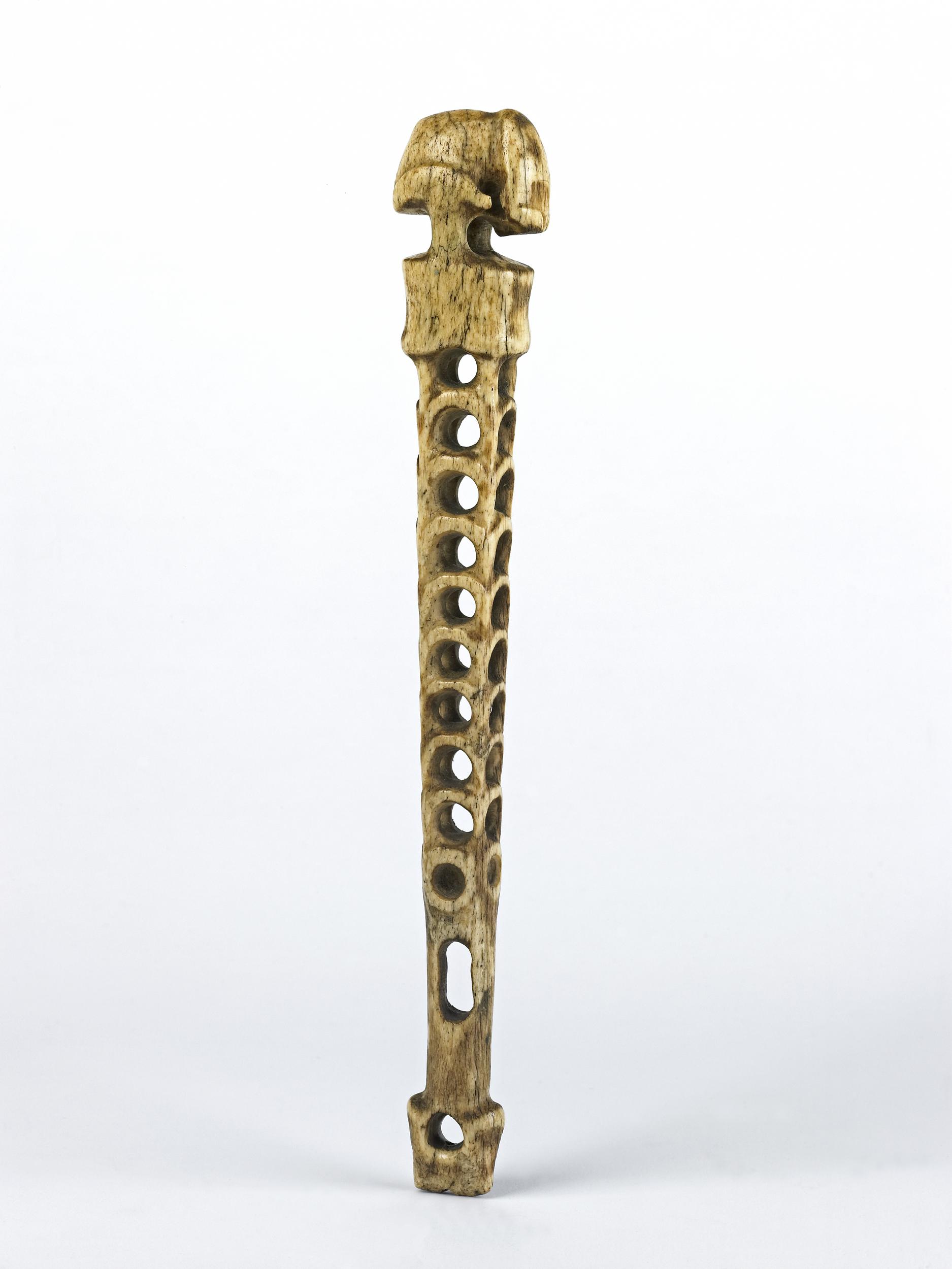 A carved bone tool with a textured top and multiple oval holes along its elongated shaft, showcasing intricate craftsmanship.