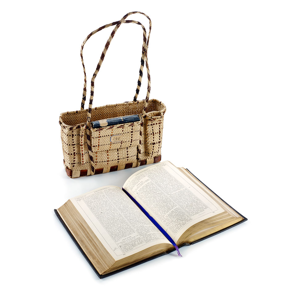 A woven straw handbag beside an open book, showcasing its textured cover and visible pages, against a white background.