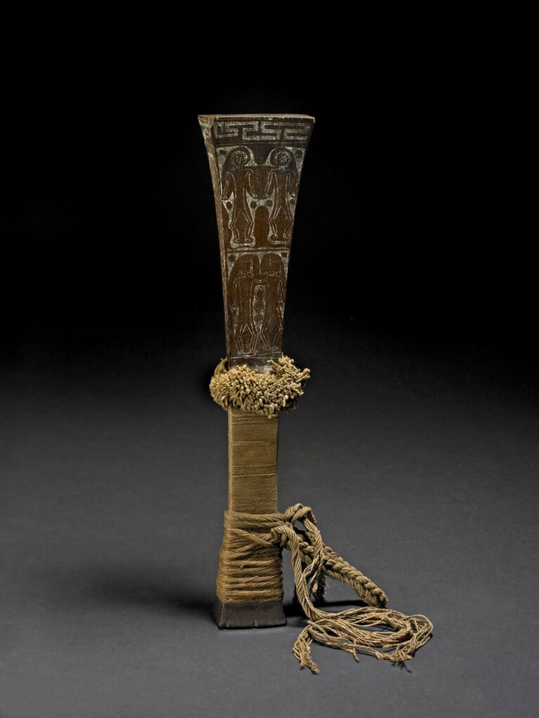 A ceremonial object featuring intricate carvings of figures, tied with textured fabric and decorative cord, against a dark background.