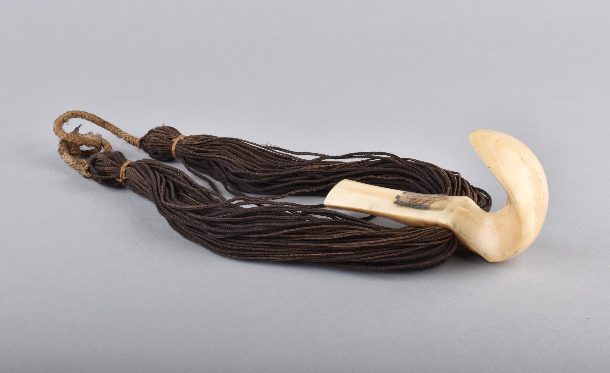 A whale tooth neck ornament, attached to long, braided dark cord, resting on a gray background.