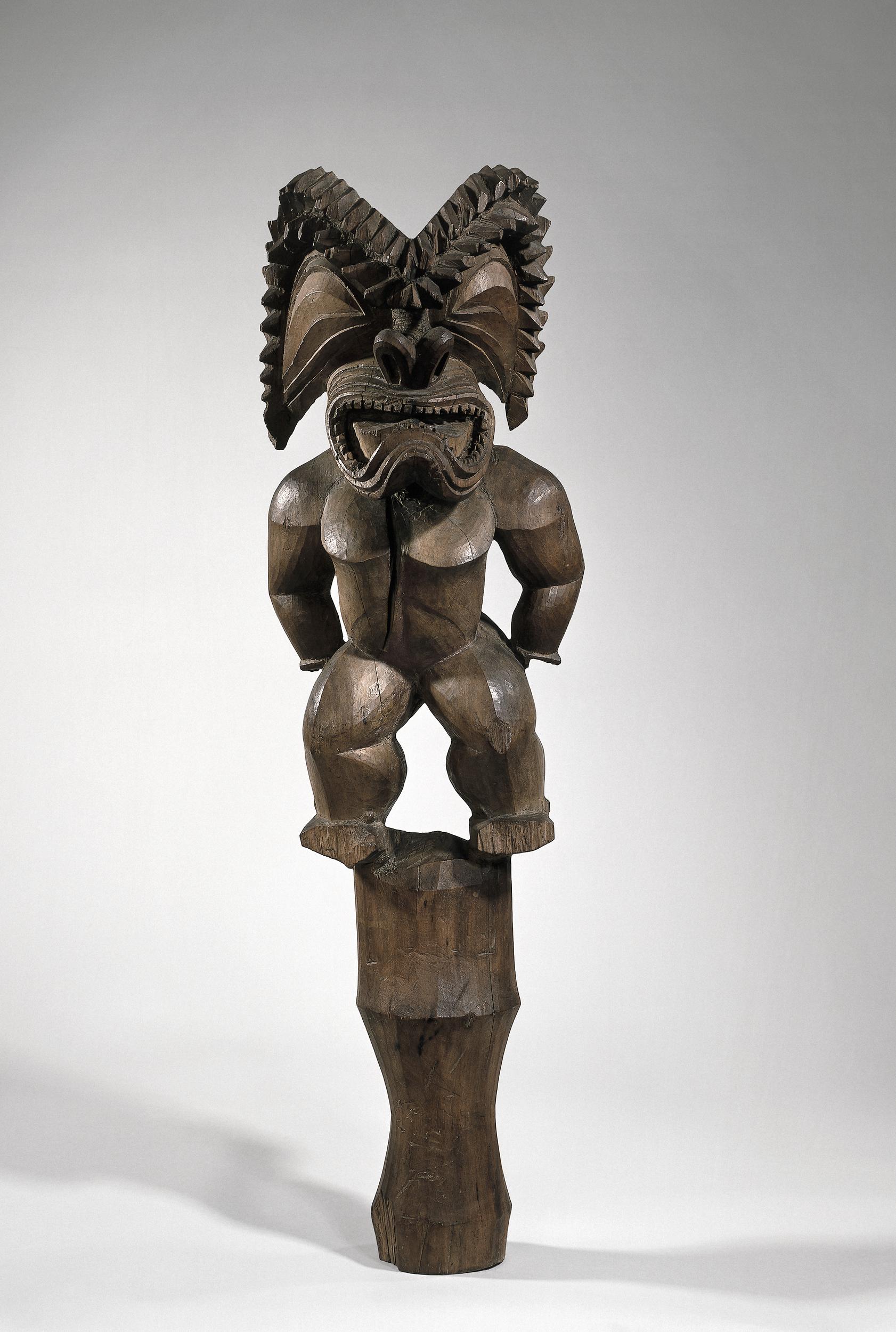 A tall, carved wooden statue depicting a muscular figure with a fierce face and elaborate headdress, showcasing intricate detailing.