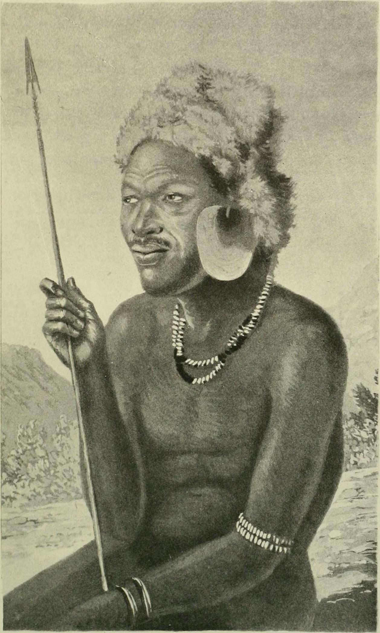 A person sits with a spear, adorned in traditional jewelry, highlighting cultural attire against a natural background.