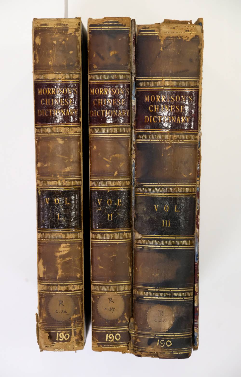 Three volumes of Morrison's Chinese Dictionary with worn leather covers and visible damage, labeled Volume I, II, and III.