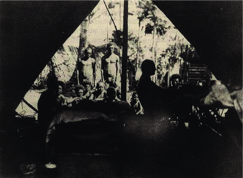 A dark, historical image captures a group of individuals seated inside a tent, with figures standing outside amidst tropical vegetation.