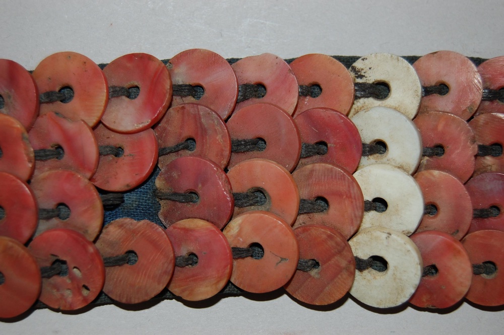 Close-up of a decorative textile featuring circular coral and shell beads, strung together with dark thread on a blue fabric base.